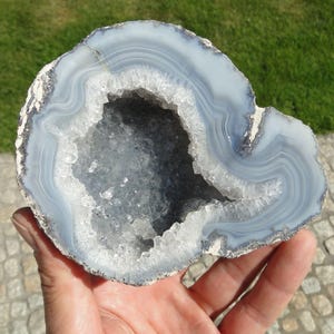 Puede incluir: Una geoda de ágata circular con bandas concéntricas de color azul claro y blanco que rodean una cavidad central llena de estructuras cristalinas translúcidas y brillantes. El borde exterior de la piedra muestra una corteza mineral rugosa.