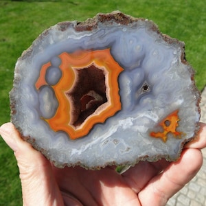Op de afbeelding: Een plak agaat met een grote, open geode in het midden. De geode is bekleed met oranje en bruine kristallen, en de omringende agaat is lichtblauw.