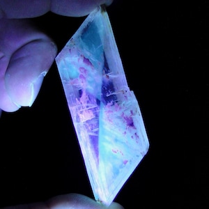 Puede incluir: Un cristal translúcido con un brillo azul y púrpura, sostenido entre los dedos sobre un fondo negro. El cristal tiene una apariencia en capas con vetas de color y una forma geométrica. El efecto general es un espécimen mineral vibrante y brillante.