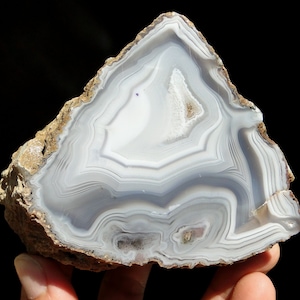 Op de afbeelding: Een grote, witte agaatgeode met een bruine buitenkant en ingewikkelde bandpatronen. De geode is doormidden gezaagd, waardoor de binnenste kristalformaties zichtbaar worden.