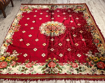 Puede incluir: Una alfombra de terciopelo rojo con un patrón floral. La alfombra tiene un medallón central con un diseño floral y un borde de motivos florales más pequeños. La alfombra tiene un borde de flecos.