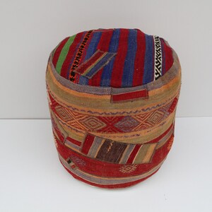 May include: A round, multi-colored, patterned ottoman with a red, blue, and green striped top. The ottoman is made of woven fabric with a variety of geometric designs.