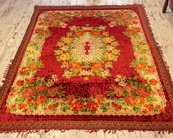 Puede incluir: Una alfombra roja con estampado floral y diseño de medallón central. La alfombra presenta un borde de flores en tonos naranja, amarillo y verde. La alfombra tiene un borde con flecos y está colocada sobre un suelo de madera.