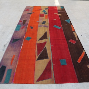 Könnte beinhalten: Ein langer, rechteckiger Teppich mit Patchwork-Muster. Der Teppich besteht aus verschiedenen farbigen Stoffstreifen, darunter Lila, Orange, Rot, Braun und Beige. Die Streifen sind zusammengenäht, um ein geometrisches Muster mit verschiedenen Formen zu bilden, darunter Dreiecke, Quadrate und Rechtecke. Der Teppich hat ein rustikales, handgewebtes Aussehen.