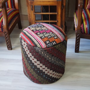 May include: A round, colorful, patterned ottoman with a woven top and a geometric design on the bottom. The ottoman is made of fabric and is sitting on a light wood floor.