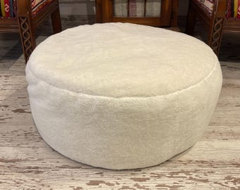 Könnte beinhalten: Ein runder, cremefarbener, plüschiger Ottoman. Die weiche, strukturierte Oberfläche ist einfarbig. Der Ottoman steht auf einem hellen, abgenutzten Holzboden. Der Hintergrund umfasst Holzstühle und einen Schrank.