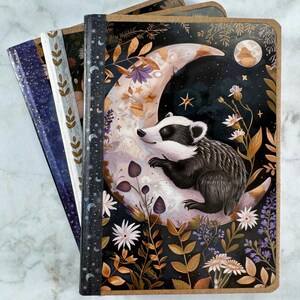 Puede incluir: Un conjunto de diarios de tapa dura con cubiertas ilustradas. Uno de los diarios presenta un tejón sobre una luna creciente con elementos florales y celestiales. Los otros diarios tienen cubiertas estampadas en tonos azules, verdes y marrones.