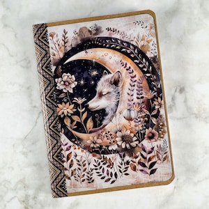 May include: A journal featuring a watercolour illustration of a fox sleeping in a crescent moon, encircled by flowers and foliage. The cover has a brown border and a patterned spine with a zig-zag design.