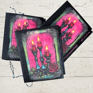 May include: A stack of black notebooks with a gothic candle design. Each notebook features a pink background with a candelabra, lit red candles, and a rose. The design is framed with a decorative border and skull accents.