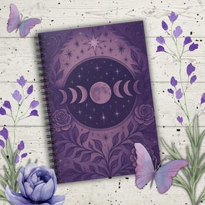 May include: A spiral-bound notebook with a purple celestial design featuring a moon phase illustration, stars, and floral accents. The cover is surrounded by lavender sprigs and butterflies, creating a mystical and artistic aesthetic.