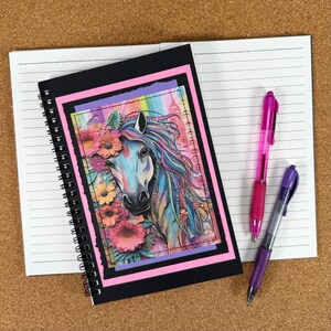 May include: A black spiral-bound notebook with a pink border, featuring a colourful illustration of a horse's face with flowers. Two pens, one pink and one purple, lie on the open, lined pages of the notebook.