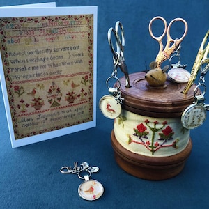 May include: A wooden spool with a mouse figurine on top, holding four pairs of scissors. The spool is decorated with a cross-stitch pattern and has a small, round, embroidered image of a bird on each side. The spool is sitting on a blue surface, next to a card with a cross-stitch pattern and a poem.