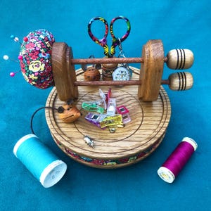 Può includere: Un portautensili da cucito in legno con un cuscino per spilli floreale, forbici colorate e una varietà di accessori per cucire. Il portautensili include una bobina di filo turchese, una bobina di filo magenta e diverse clip da cucito colorate. È presente anche una piccola statuina di topo.