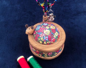 May include: A wooden pincushion with a floral print and a mouse figurine on the side. The pincushion has a built-in scissor holder with a pair of black and white scissors. Two spools of thread, one red and one green, are in the foreground.