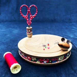 Può includere: Un kit da cucito in legno con una forbice a pois rossi e bianchi, un cuscinetto per spilli a forma di topo e un'bobina di filo rosso. Il kit ha un design floreale attorno al bordo.