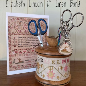Può includere: Un telaio da ricamo in legno con un nastro di lino da 5 cm, che presenta un disegno a punto croce con un motivo floreale con fiori rosa e gialli. Il telaio è pieno di forbici da ricamo, un piccolo topo di legno e un ciondolo con un cervo.