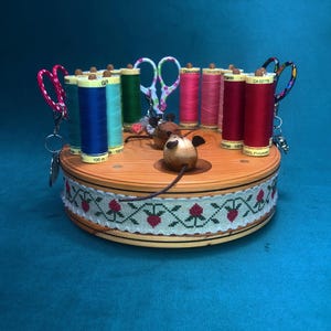 May include: A round wooden sewing organizer featuring a light-colored wood grain base wrapped in a cream-colored ribbon with a red strawberry pattern. It holds multiple spools of colorful thread, two pairs of small scissors, and two wooden mouse figures.