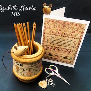 May include: A wooden needle holder with a mouse figurine on top, filled with knitting needles. A card with a sampler featuring the alphabet and a poem by Elizabeth Lincoln, dated 1815, is propped against the holder. A pair of scissors and a charm with a deer and a heart are also visible.
