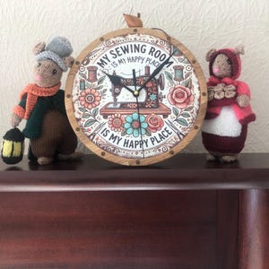 Op de afbeelding: Een ronde houten klok met de tekst "MY SEWING ROOM IS MY HAPPY PLACE" en een illustratie van een naaimachine. Twee gebreide muisfiguren flankeren de klok. Eén muis houdt een lantaarn vast.