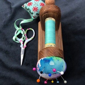 May include: A wooden sewing thread holder with a spool of light blue thread, a small green and red cross-stitch cushion, a pair of pink and white scissors, and a blue and white pincushion with colourful pins.