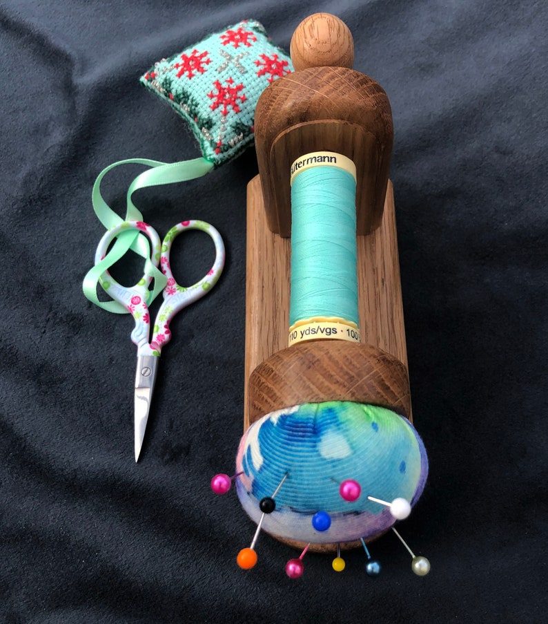 May include: A wooden sewing thread holder with a spool of light blue thread, a small green and red cross-stitch cushion, a pair of pink and white scissors, and a blue and white pincushion with colourful pins.