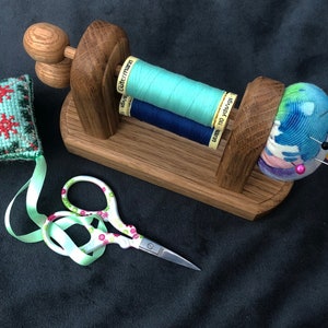 May include: A wooden sewing thread holder with three spools of thread, a pincushion with colourful pins, a small green and red embroidered cushion, and a pair of scissors with a floral design.