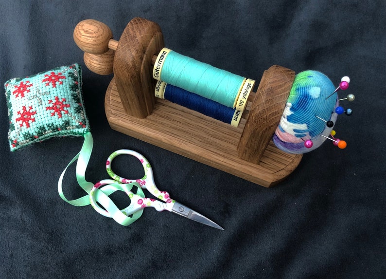 May include: A wooden sewing thread holder with three spools of thread, a pincushion with colourful pins, a small green and red embroidered cushion, and a pair of scissors with a floral design.