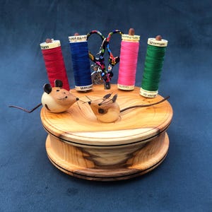 Può includere: Un organizer da cucito in legno dal design circolare, con due topolini in legno intagliato e rocchetti di filo rosso, blu, rosa e verde. Un paio di forbici con motivi floreali sono al centro. L'organizer è in legno chiaro.