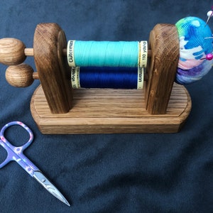 May include: Wooden thread holder with two spools of thread, one blue and one light blue, and a pincushion with a colourful fabric cover. The pincushion has several pins stuck in it.