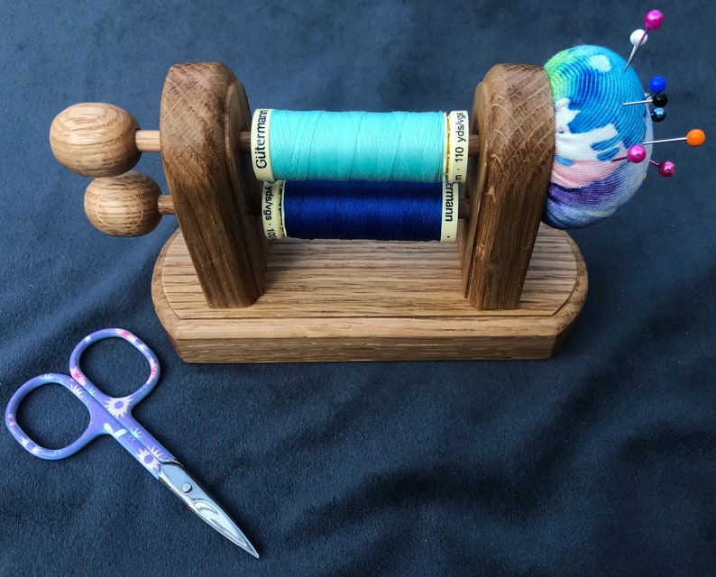 May include: Wooden thread holder with two spools of thread, one blue and one light blue, and a pincushion with a colourful fabric cover. The pincushion has several pins stuck in it.