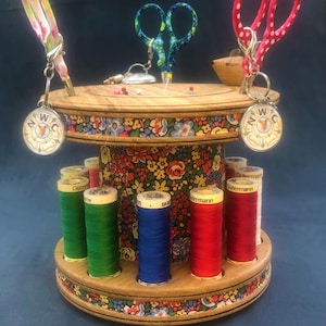 May include: A wooden sewing organiser with floral trim and a floral fabric centre. It holds spools of colourful thread, decorative scissors, and pins. The organiser has a vintage aesthetic.