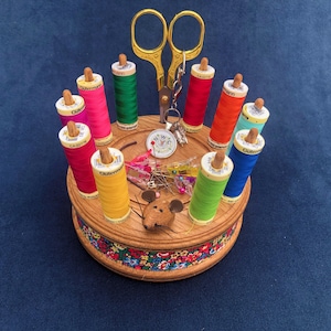Può includere: Un porta-filo di legno con un design a forma di topo. Ha una base circolare con un motivo floreale e contiene otto bobine di filo di vari colori, tra cui rosso, rosa, giallo, verde, blu e arancione. Sono inclusi anche un paio di forbici dorate e una piccola bussola.
