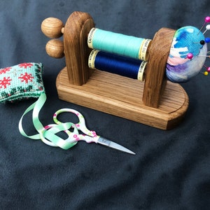 May include: Wooden sewing needle holder with two spools of thread, one blue and one green, and a pincushion with colourful fabric and pins.