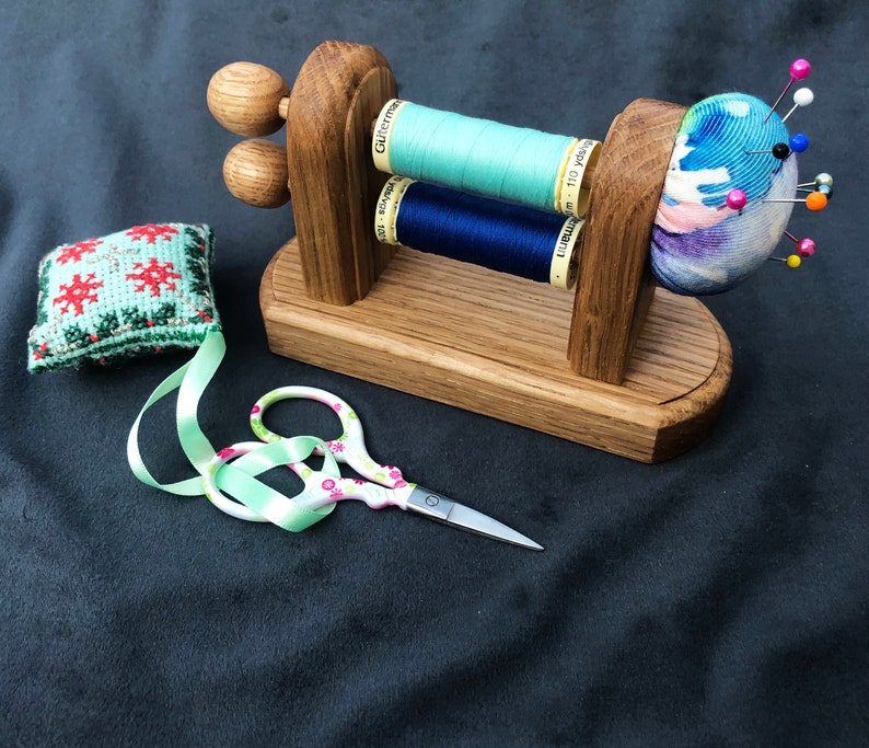 May include: Wooden sewing needle holder with two spools of thread, one blue and one green, and a pincushion with colourful fabric and pins.