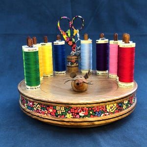 Può includere: Un organizer da cucito in legno con rocchetti di filo colorati, forbici floreali e una statuina di topo in legno. L'organizer ha un motivo floreale attorno alla base e contiene rocchetti di filo verde, giallo, blu, azzurro, viola, rosa e rosso.
