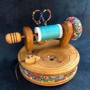 May include: A wooden sewing organiser featuring a circular base with a floral trim, a spool holder with turquoise thread, a floral pincushion, a pair of patterned scissors, and a small wooden mouse figurine with three sewing pins on the base.