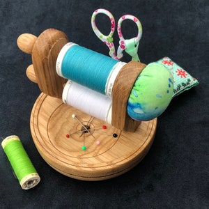 Può includere: Un supporto in legno per rocchetti di filo con due rocchetti di filo, uno blu e uno bianco, un cuscino per spilli e un paio di forbici rosa e verdi. Il supporto ha una base circolare con un cuscino per spilli a forma di stella al centro.