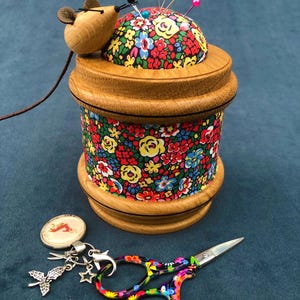 Può includere: Un cuscino per spilli in legno con un rivestimento in tessuto floreale e un coperchio a forma di topo. Il cuscino per spilli è pieno di spilli e si trova su una base in legno. Sono anche raffigurati un paio di forbici colorate e un ciondolo con una farfalla e stelle.