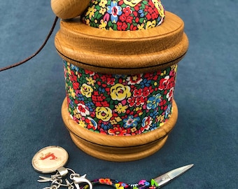 May include: A wooden pincushion with a floral fabric cover and a mouse-shaped lid. The pincushion is filled with pins and sits on a wooden base. A pair of colourful scissors and a charm with a butterfly and stars are also shown.