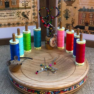 Może przedstawiać: Drewniany stojak do szycia z kolorową, kwiatową obwódką. Stojak ma małą figurkę myszy, kolekcję kolorowych szpulek nici, parę nożyczek i poduszkę do szpilek ze szpilkami.