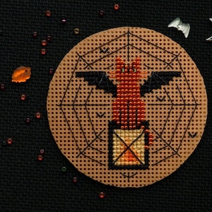 May include: A cross-stitch pattern featuring a red cat with bat wings sitting on a box with a spiderweb design. The pattern is on a brown background with a white border.
