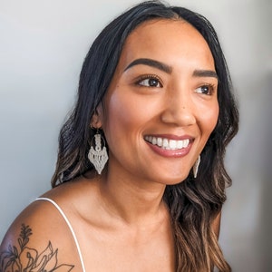May include: A woman with long brown hair is wearing a white tank top and a pair of white macrame earrings. She is smiling and looking to the right.