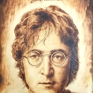 May include: A woodburned portrait of John Lennon with his signature and the word "Imagine" above his head. The portrait is in shades of brown and white.