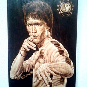Puede incluir: Un retrato de Bruce Lee quemado en madera, en una postura de lucha, con un símbolo yin yang y caracteres chinos en el fondo.