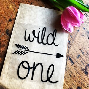Könnte beinhalten: Ein schwarzer Vinyl-Aufkleber mit den Worten "wild one" in kursivschrift. Ein Pfeil befindet sich zwischen den Wörtern. Der Aufkleber befindet sich auf einem braunen Papierhintergrund.