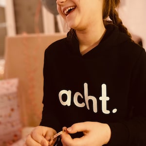 Op de afbeelding: Een jong persoon draagt een zwarte hoodie met het woord "acht" in wit op de voorkant. De persoon lacht en houdt een zilveren armband vast.