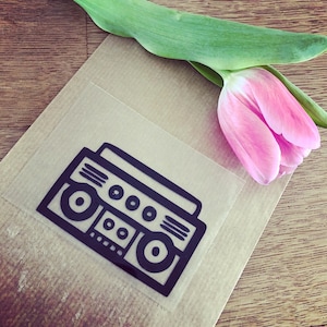 Op de afbeelding: Een zwart-witte illustratie van een vintage boombox op een bruine papieren achtergrond. Op de achtergrond is een roze tulp te zien.