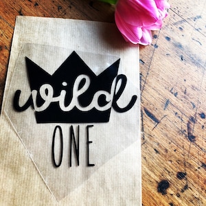 Op de afbeelding: Een zwart kroonontwerp met de woorden "wild one" in een scriptlettertype en "one" in een schreefloos lettertype. Het ontwerp staat op een transparante achtergrond, geplaatst op bruin papier. Een roze tulp staat in de rechterbovenhoek.