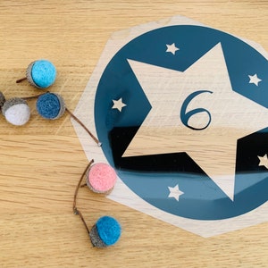 Könnte beinhalten: Ein blauer und weißer Stern mit der Nummer 6 in der Mitte. Der Stern befindet sich auf einem durchsichtigen Aufkleber mit blauem Hintergrund und weißen Sternen. Auf einer Holzoberfläche liegen mehrere kleine Filzkugeln in verschiedenen Farben.