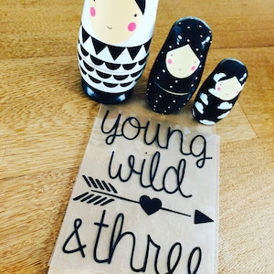 Könnte beinhalten: Ein Set aus drei schwarzen und weißen Nistpuppen mit einem schlichten, modernen Design. Die größte Puppe hat ein schwarz-weißes geometrisches Muster, die mittlere Puppe hat schwarze Punkte und die kleinste Puppe hat ein schwarz-weißes Wolkenmuster. Die Puppen stehen auf einer braunen Oberfläche mit einem Aufkleber, auf dem "young wild & three" steht.